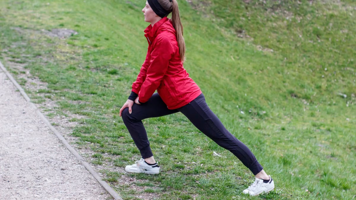 Woman in red jacket stretching in a park, illustrating outdoor fitness and healthy lifestyle.